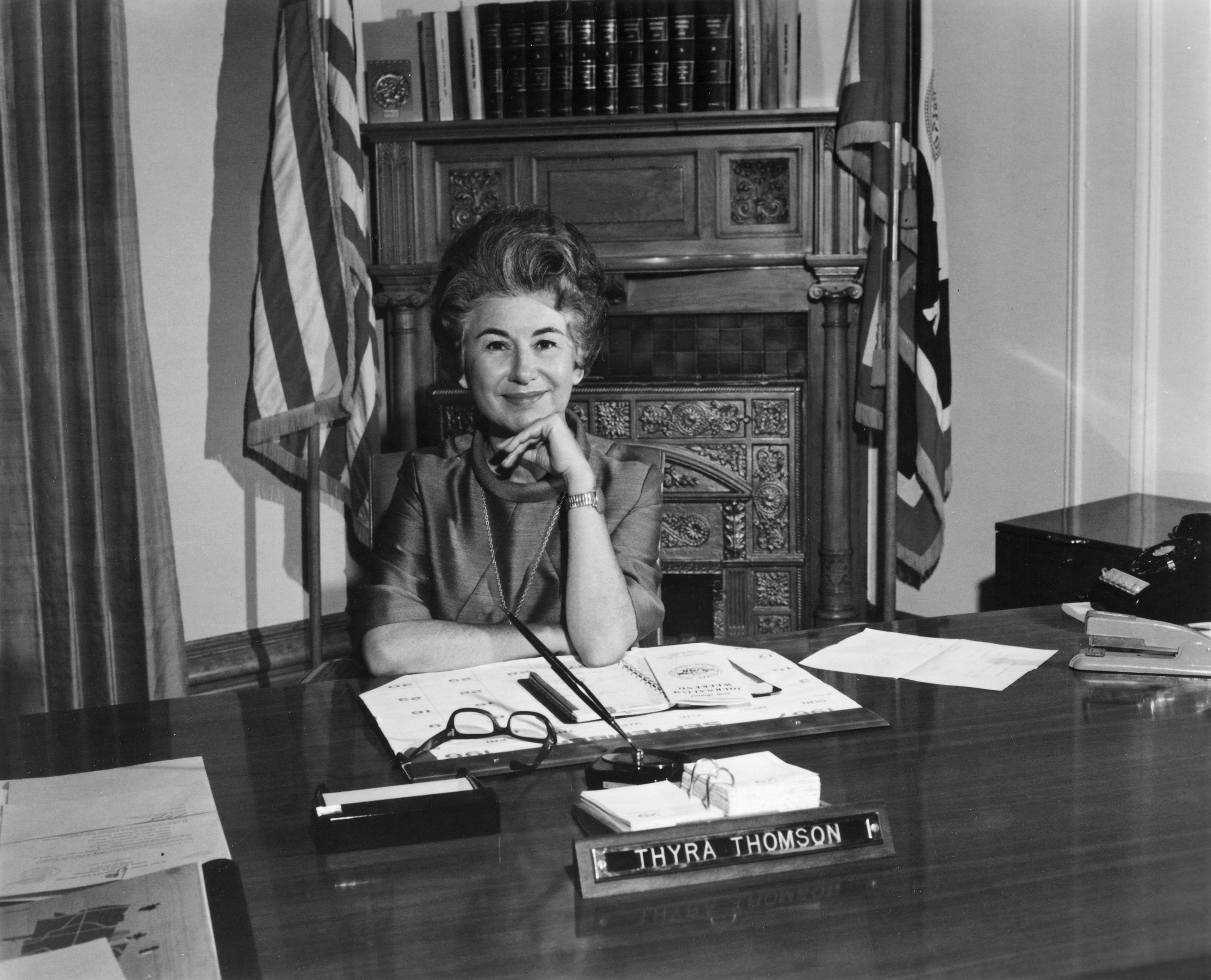 woman sitting at a large, official looking desk with american flags behind her